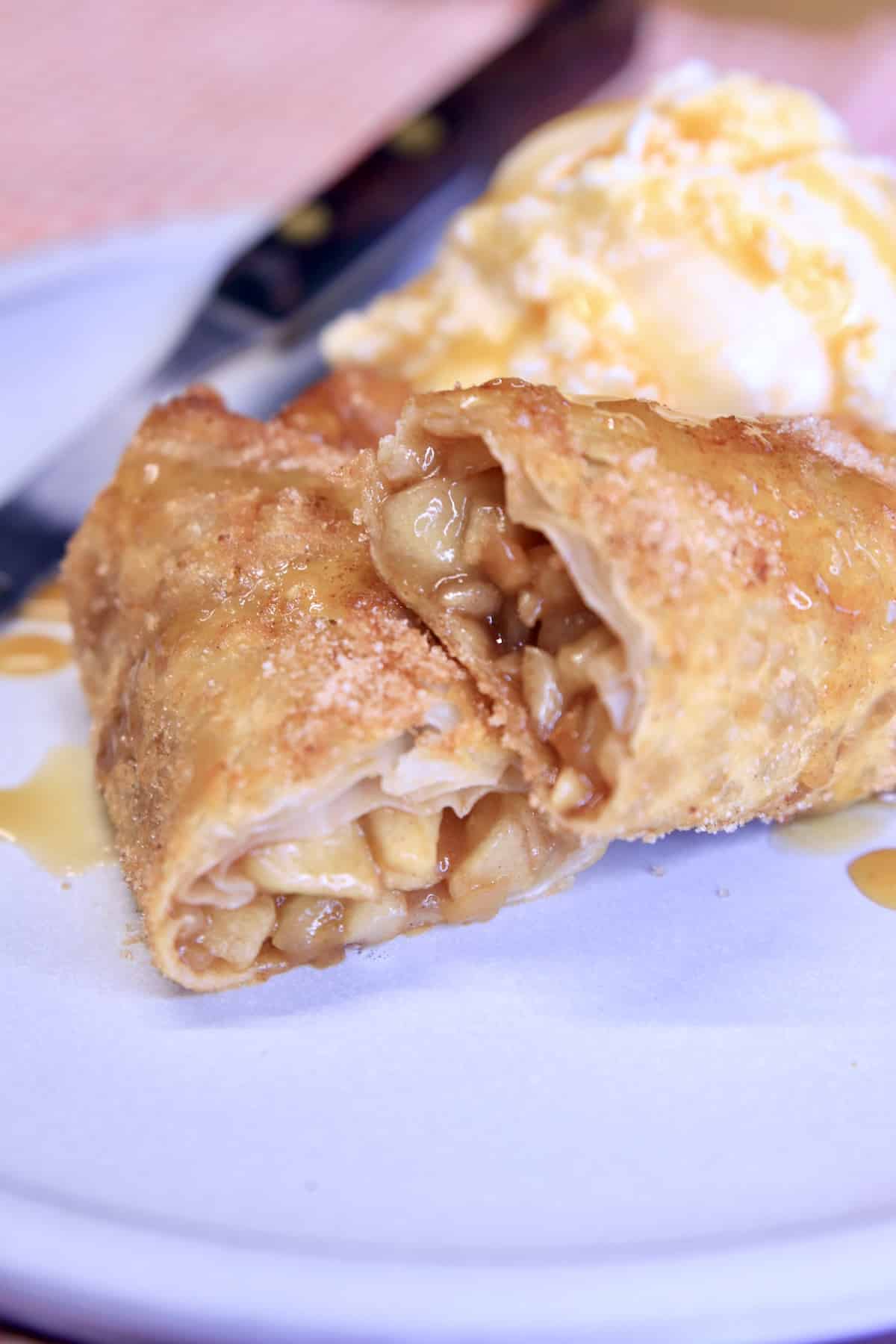 Apple Pie Egg Roll sliced in a half on plate with ice cream and caramel sauce.