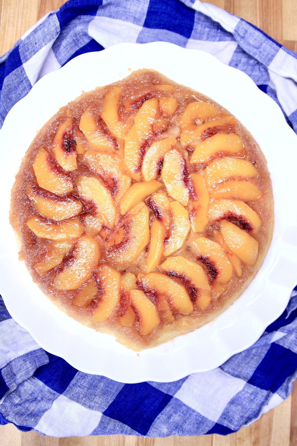 Peach upside down cake on a white platter, blue and white check cloth beneath.