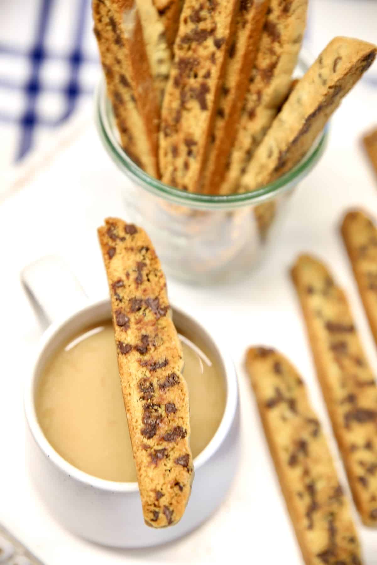 Chocolate chip biscotti on a coffee cup, in a jar, on a white board.