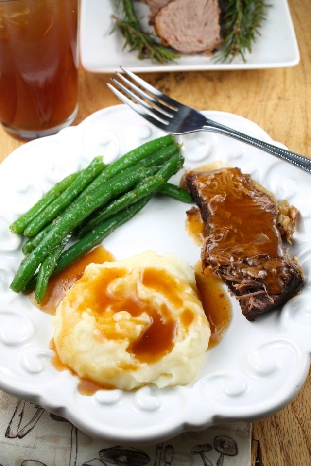 Pot Roast slice with gravy, mashed potatoes topped with gravy and green beans on a plate with a glass of tea