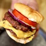 Hand holding double cheeseburger with tomato and red onion.