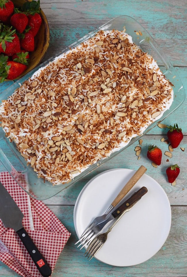Coconut Cream Cake topped with toasted coconut in a glass baking dish, white plates with forks, whole strawberries and a cake knife on a red check napkin