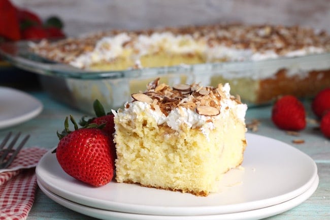 Slice of easy Coconut Cream Cake on a plate with whole strawberries with cake pan in the background