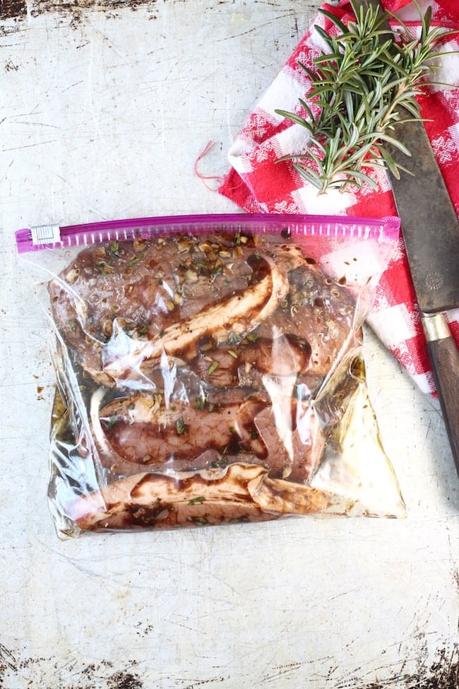 Zip top bag with pork chops in marinade, red napkin, fresh rosemary and a knife on baking sheet
