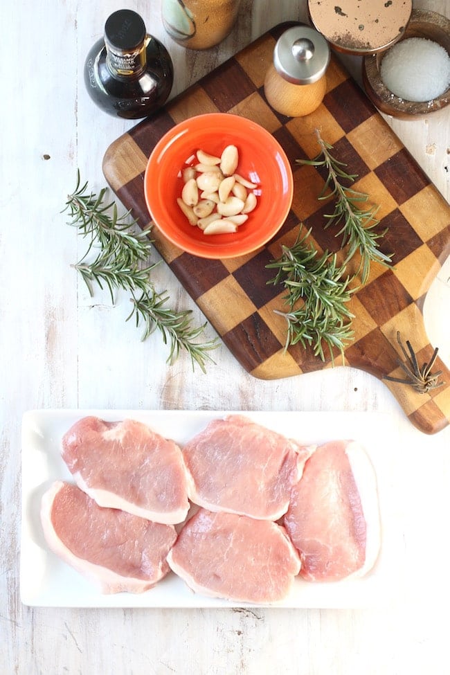 Boneless Pork Chops, peeled garlic cloves in an orange bow, fresh rosemary on a checkered cutting board, olive oil carafe, salt cellar, pepper grinder on a white back ground