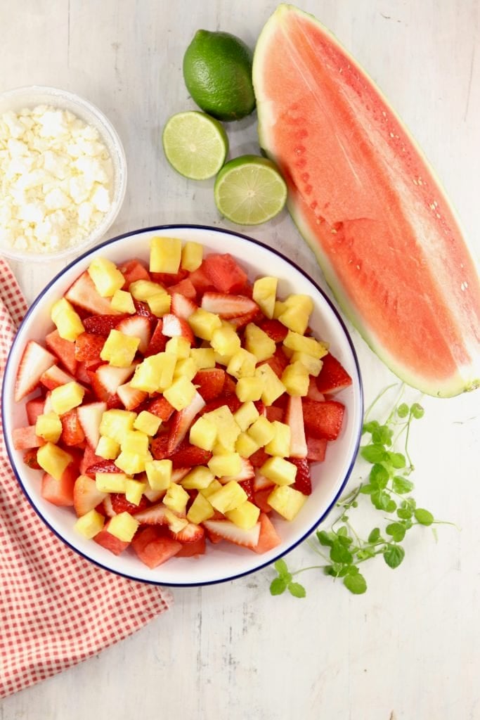 watermelon, strawberries, and pineapple in a bowl plus a bowl of feta cheese, limes and watermelon wedge
