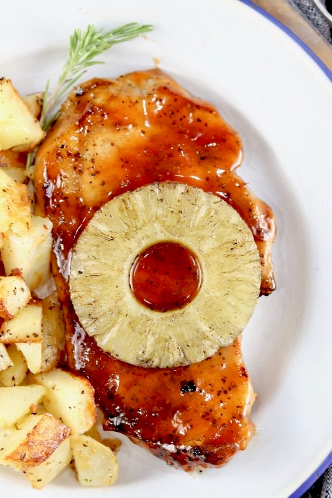 Grilled BBQ Pork Chops topped with a slice of pineapple and potatoes on the side on a white plate