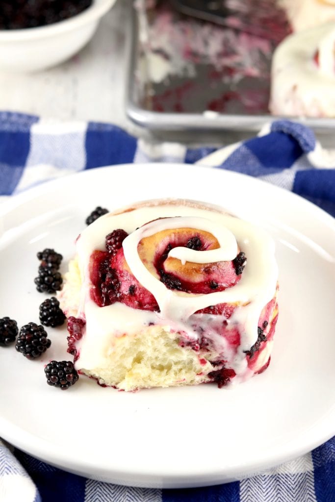 White plate with sweet roll filled with blackberry filling and topped with cream cheese icing in a swirl. 