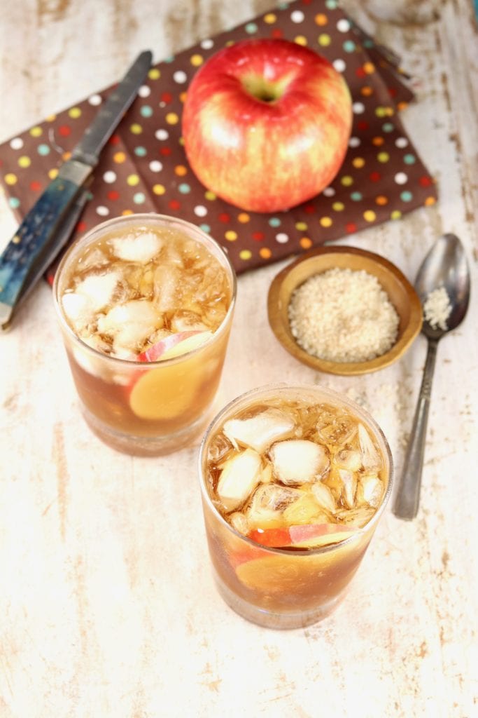 2 glasses of spiked apple cider with a fresh apple and small bowl of vanilla sugar