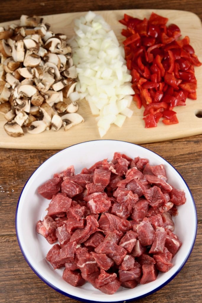 Cubed sirloin steak in a bowl, cutting board with chopped mushrooms, onions and red bell peppers