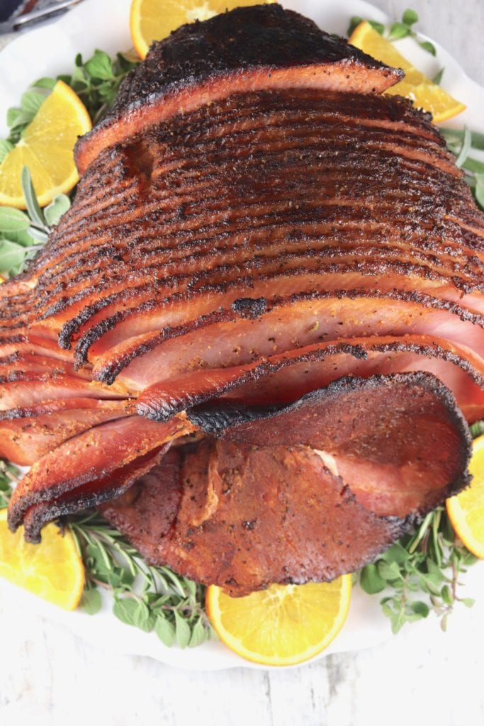 Top view of spiral sliced ham with maple glaze on a platter with herbs and sliced oranges