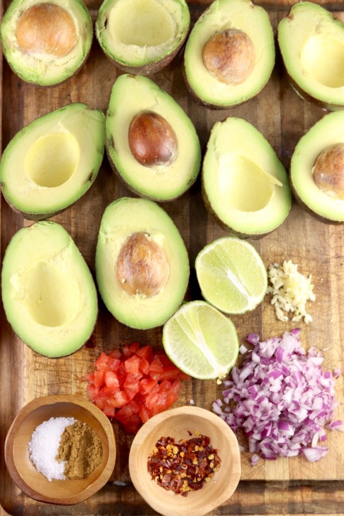 Ingredients for guacamole, avocados cut in half, red onion, garlic, tomatoes, salt, red pepper flakes