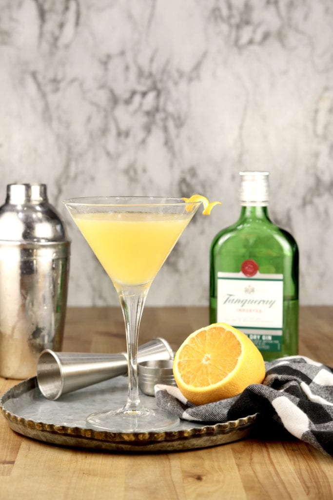 Bees Knees Cocktail in a Martini Glass with a lemon twist, Bottle of Tanqueray Gin, half of a lemon and a cocktail shaker on a tray