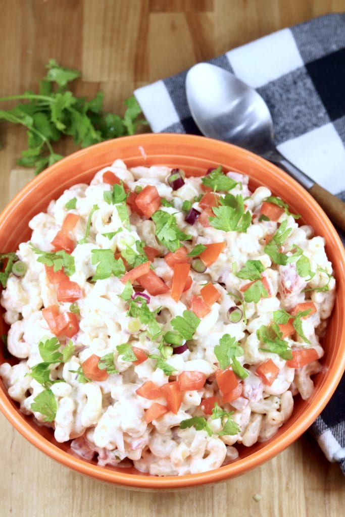 Macaroni salad with Mexican flavors, tomatoes, onions, jalapeno, cilantro in an orange bowl. Patterned napkin with a spoon off to the side