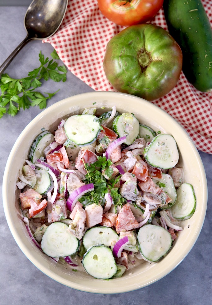 Creamy cucumber tomato salad in a bowl, fresh tomatoes, herbs and cucumbers along side with serving spoon and red napkin