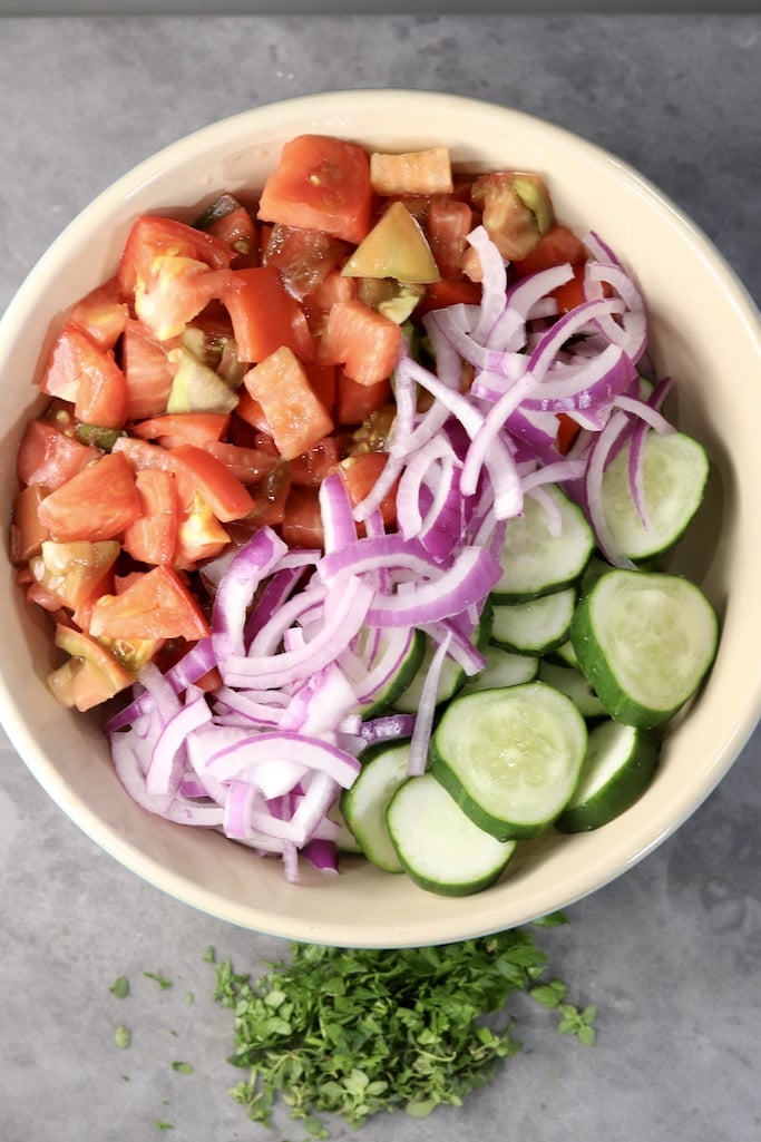 Cut up tomatoes, red onions sliced and sliced cucumbers in a bowl, fresh herbs below