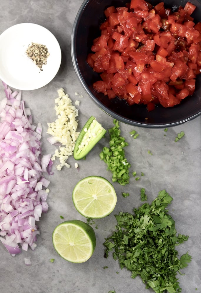 diced tomatoes in a bowl, diced red onion, garlic, cliantro. lime halves and sliced jalapeno on a cutting board with a pinch bowl of salt and pepper