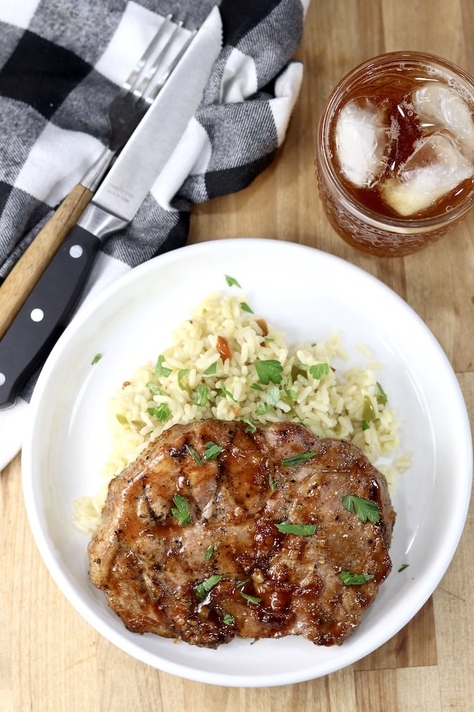Plate with rice pilaf and grilled peach bbq pork steaks, black and white napkin, fork, knife and glass of tea around the plate