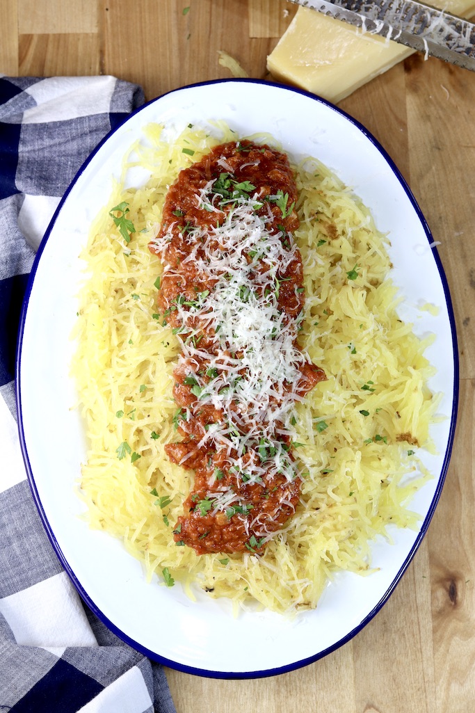 Roasted tomato sauce over spaghetti squash on an oval platter, topped with green herbs and fresh grated parmesan cheese. Wedge of parmesan near the top and black napkin to the side