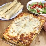 Pan of baked lasagna, platter of breadsticks and bowl of salad topped with tomatoes