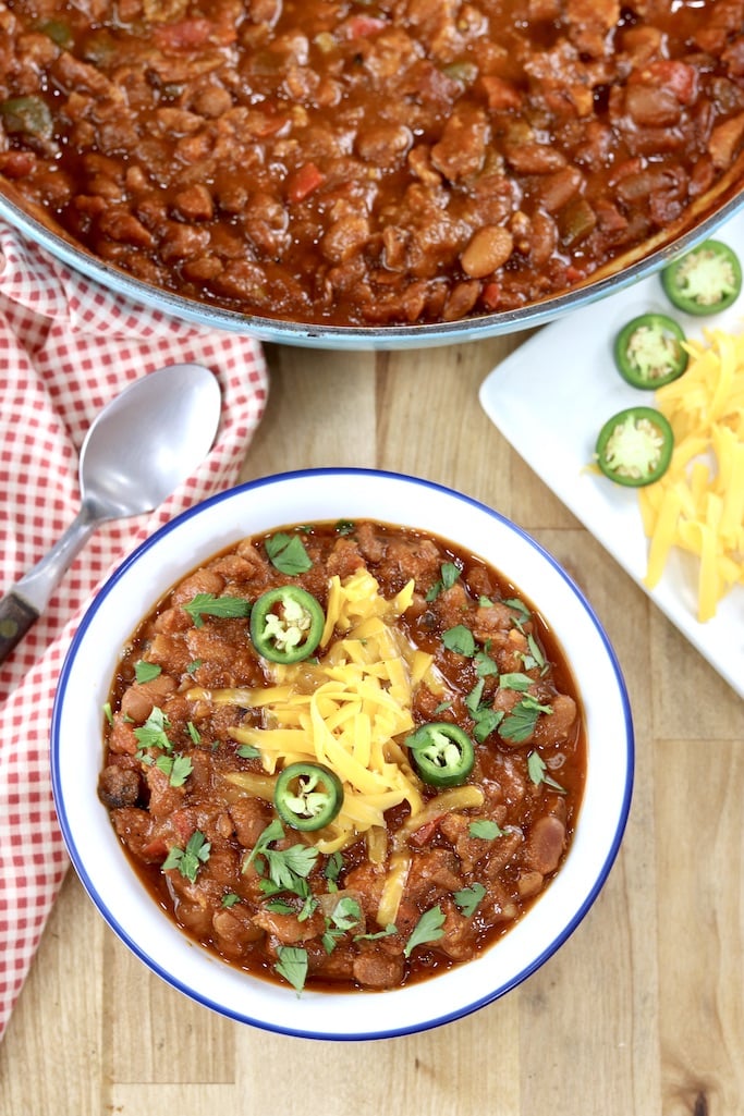 Bowl of chili with cheese, jalapenos, pan of chili above on a wood table