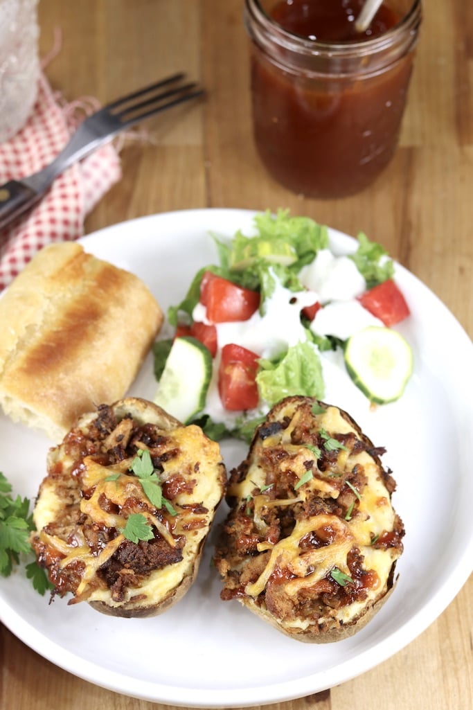 Twice baked potatoes topped with pork, served with salad and bread on a white plate. BBQ sauce jar in background.