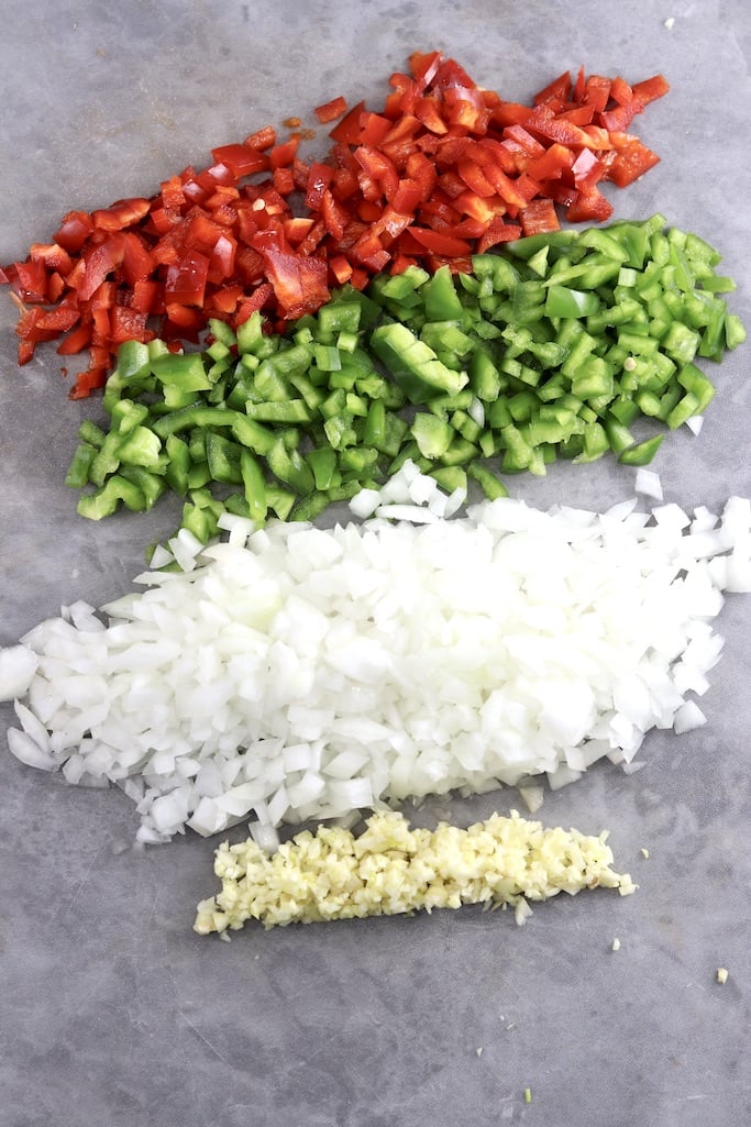 diced red bell peppers, green bell peppers, onions and garlic on a gray cutting board
