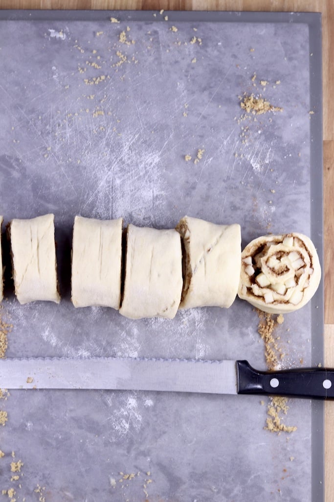 Sliced cinnamon roll dough with serrated knife below on gray board