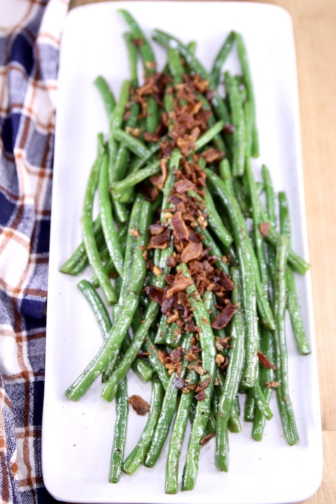 Rectangle white platter with cooked green beans topped with crispy crumbled bacon