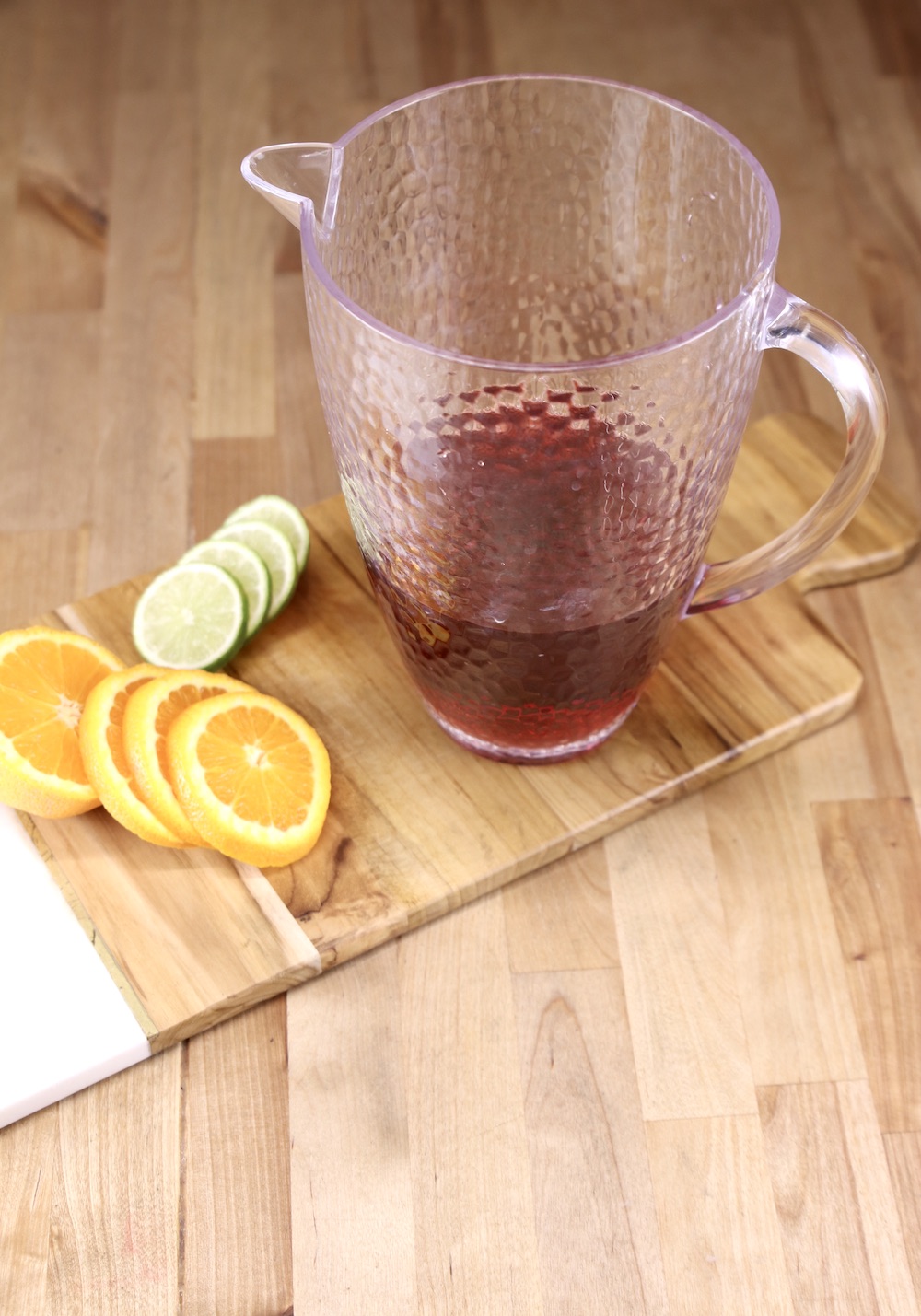Pitcher with cranberry juice on a wood board with sliced oranges and limes