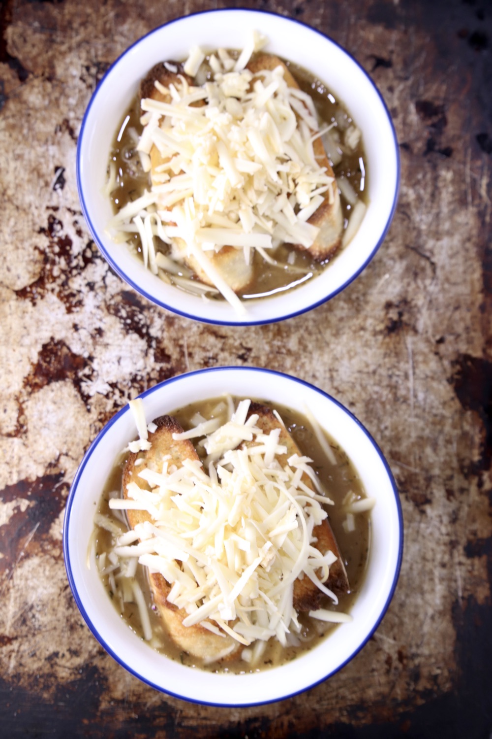 2 bowls of French Onion Soup on a baking sheet topped with toasted baguette and shredded cheese