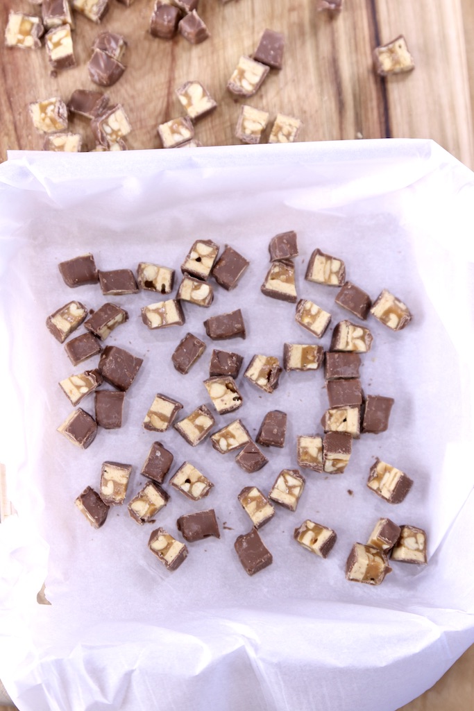 square dish lined with parchment paper, with chopped snickers bars
