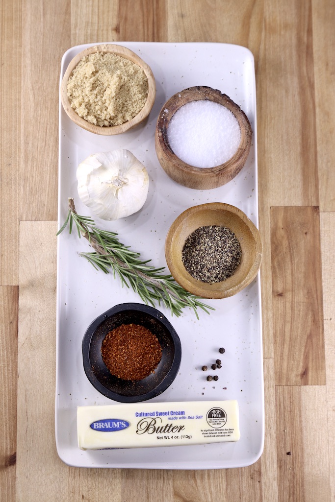 Ingredients for beef tenderloin garlic butter crust on a white plate: butter, red pepper, black pepper, salt, head of garlic, brown sugar and sprig of rosemary