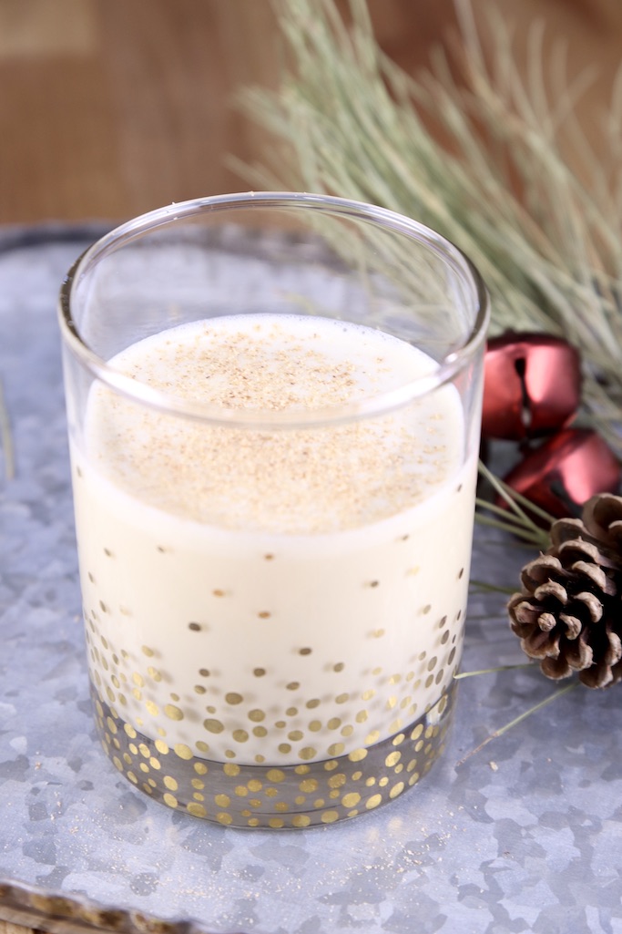 Glass of spiked eggnog on a tray with pine branch, pine cone and red bells