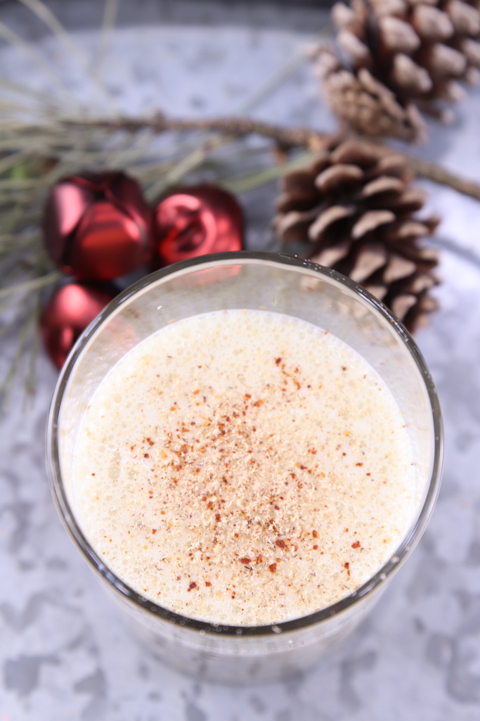 Spiked Eggnog cocktail with nutmeg and red pepper garnish. Galvanized tray background with pinecones and red jingle bells