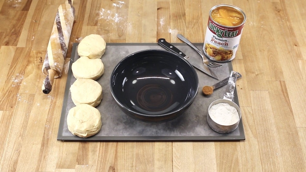 Cutting board with can biscuits, a black bowl, rolling pin, can of peach pie filling, spoon of cinnamon, flour