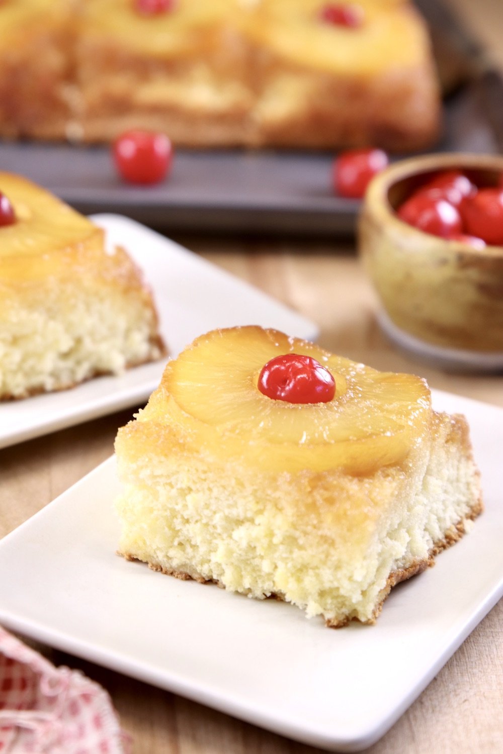 Slice of pineapple upside down cake on a small plate, bowl of cherries, platter of cake in background