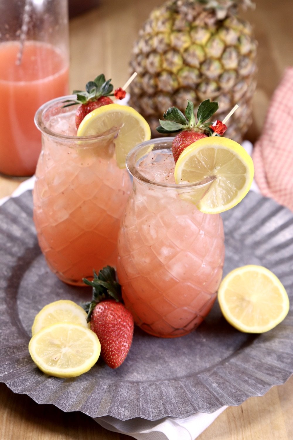 2 tiki glasses with strawberry lemonade on a platter with lemons, strawberries