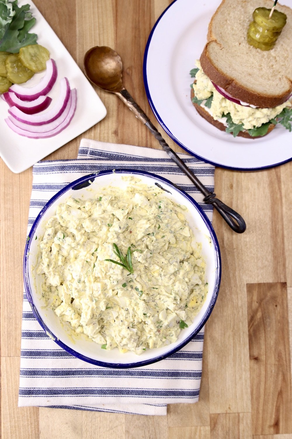 Bowl of egg salad, plate with red onions, lettuce and sandwich