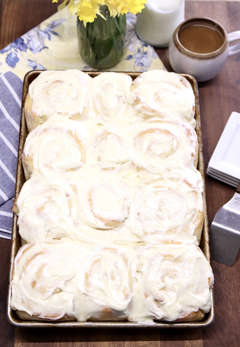 pan of cream cheese iced sweet rolls , flowers and coffee cup in background