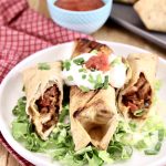 short rib burritos on a plate - one whole, one cut in half, with shredded lettuce, sour cream and salsa