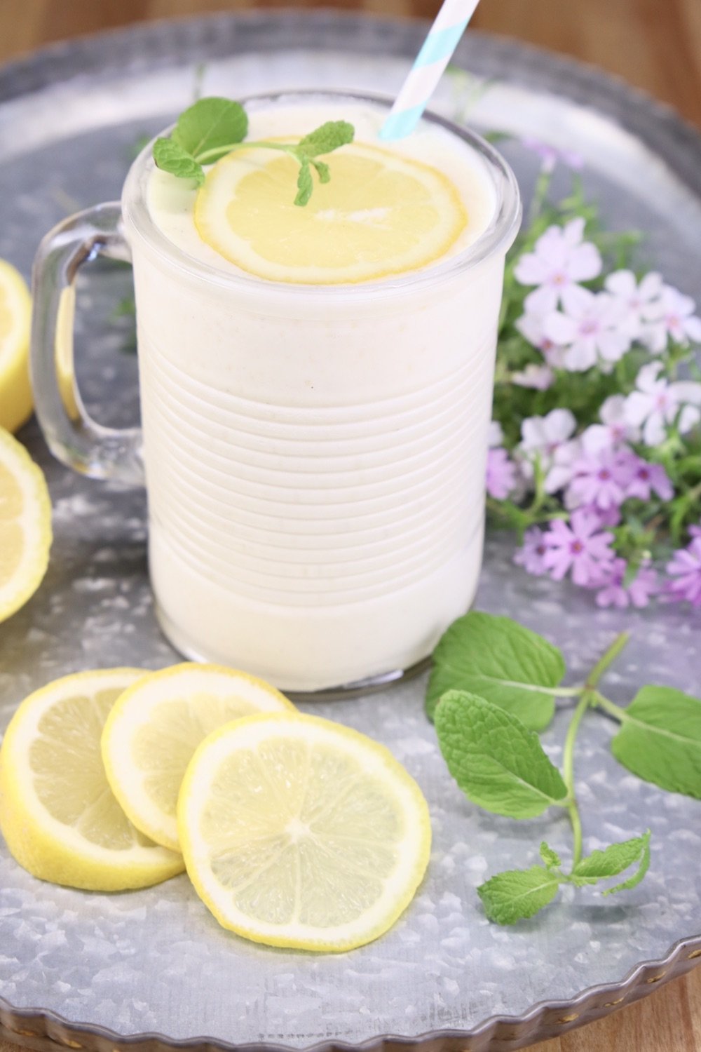 Frosted pineapple lemonade in a glass with lemon and a straw. Sitting on a tray with lemon slices, fresh flowers
