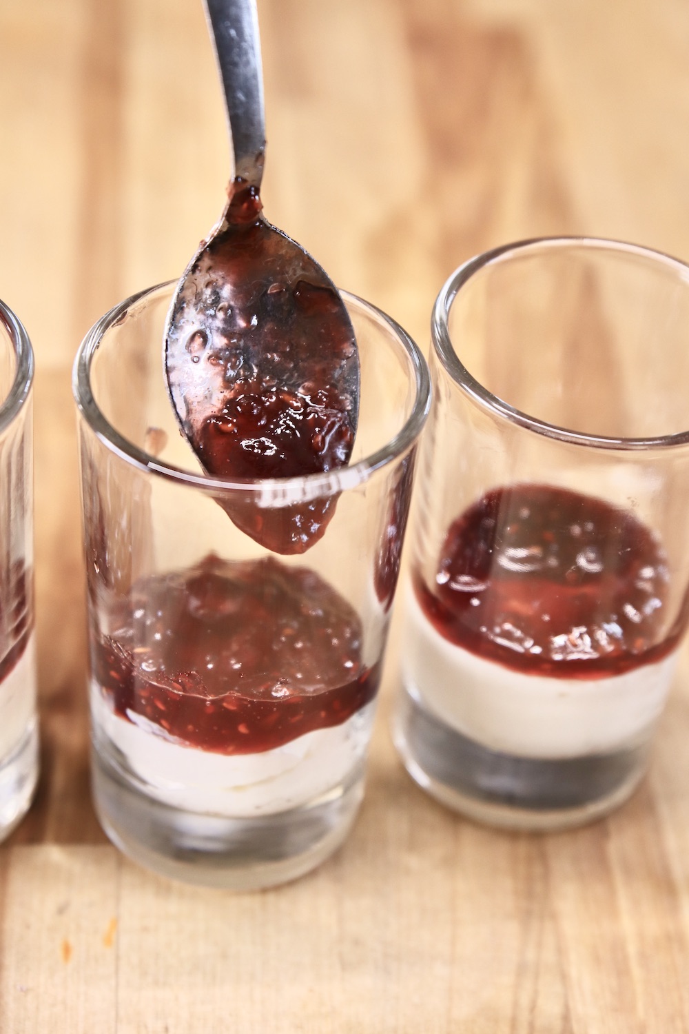 spooning raspberry preserves into a dessert glass with cheesecake filling
