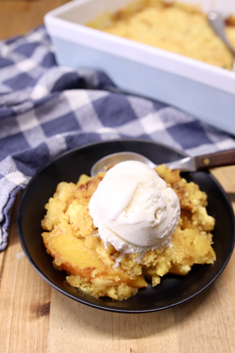 Peach Dump Cake with vanilla ice cream in a black bowl, cake pan in background