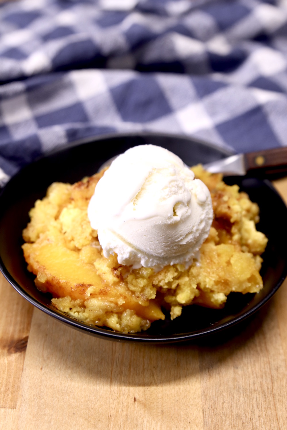 Peach Dump Cake in a black bowl with vanilla ice cream - blue towel in background