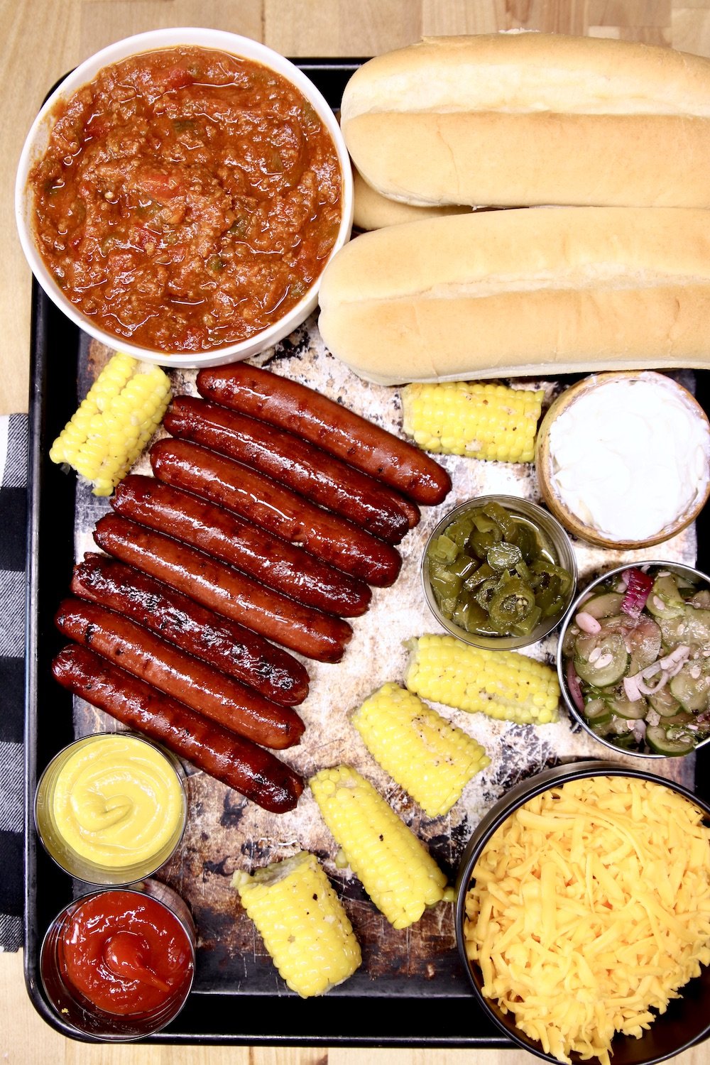 Baking sheet with hot dog chili, buns, grilled hot dogs, corn on the cob, shredded cheese, condiments