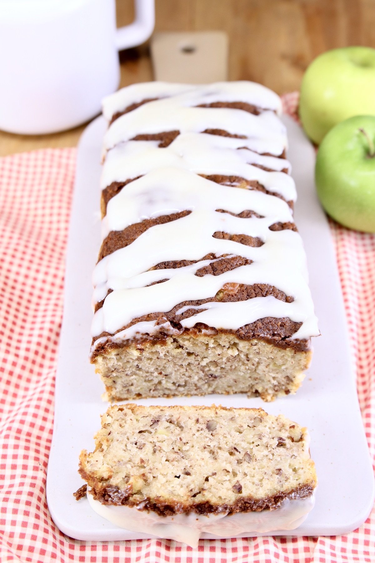 Apple bread with cream cheese drizzle on a board - one slice in front of loaf, apples in back ground