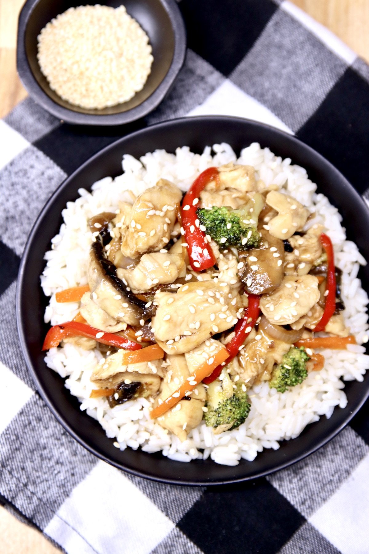 chicken stir fry with sesame seeds over rice in a black bowl, small bowl of sesame seeds