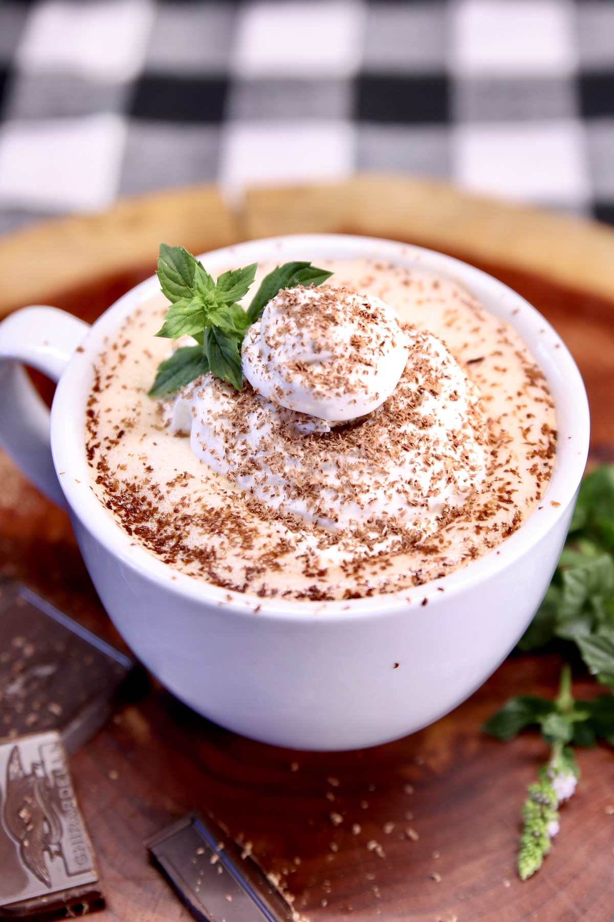 mug of cocoa topped with whipped cream, shaved chocolate and mint sprig on a wood board