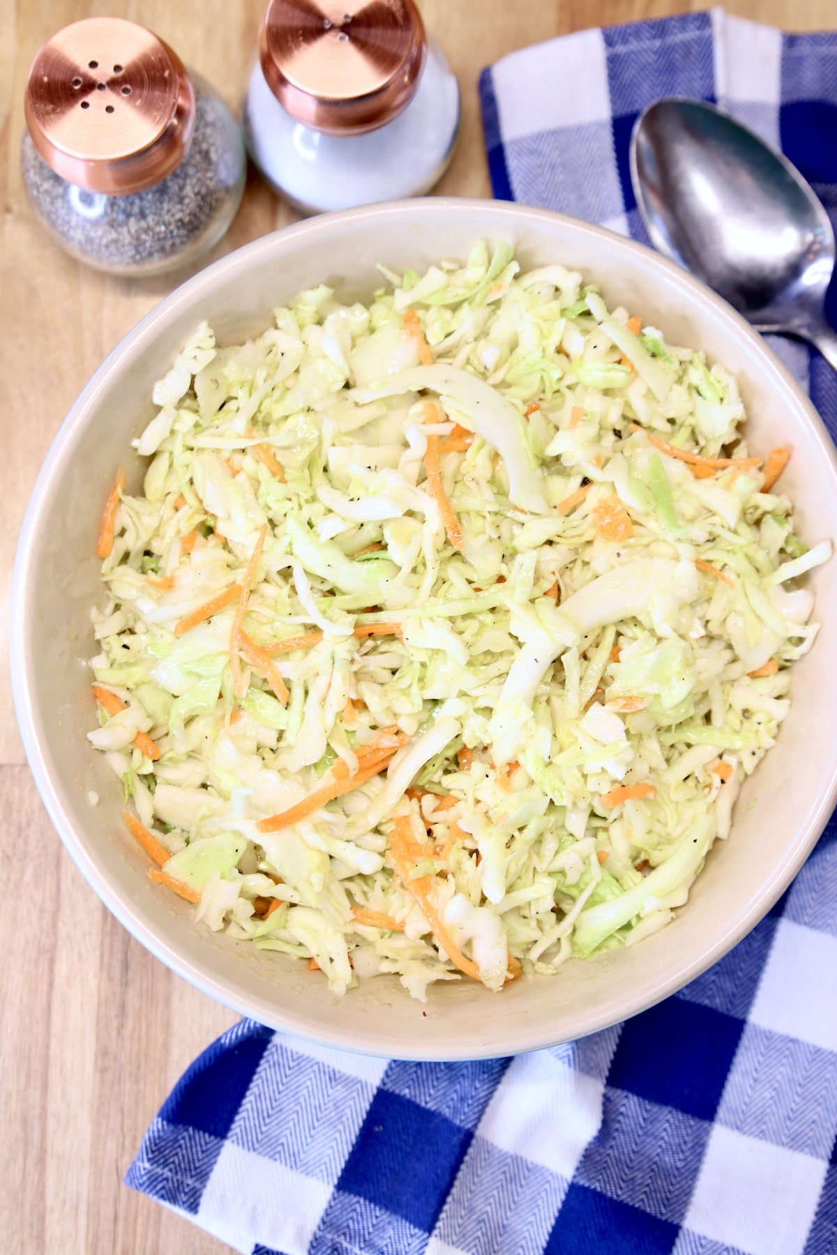 overhead of bowl of coleslaw on a blue check napkin. Salt & pepper shakers to the side.